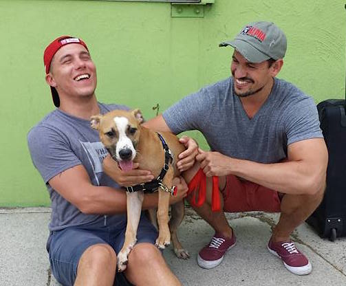 A photo of two men sitting on the sidewalk outside of Liberty Humane Society while their newly adopted dog Yandi licks their faces.